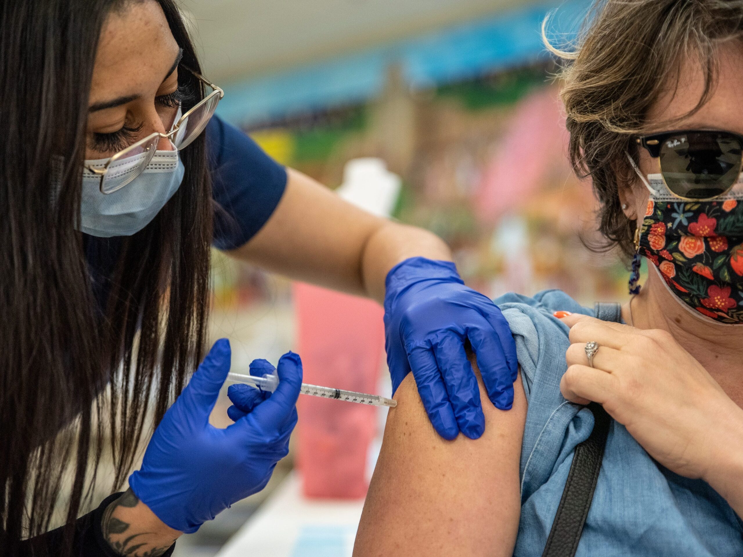 Women with mask and glasses administers COVID-19 vaccine to middle-aged women with sunglasses and patterned mask