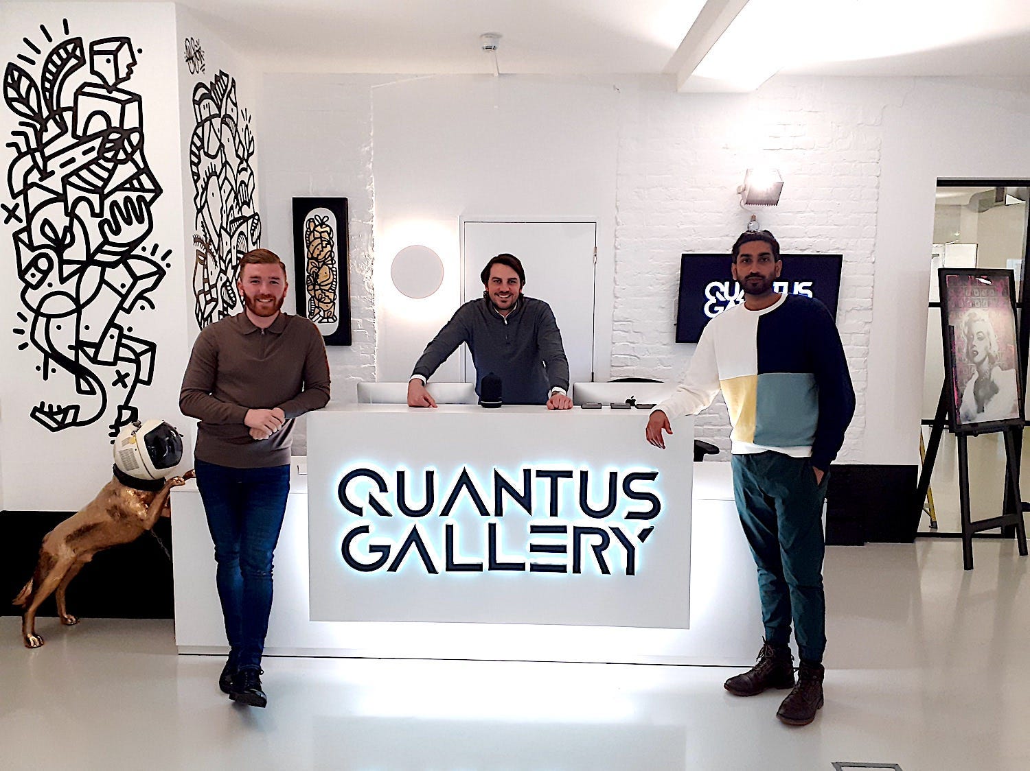 The three founders pose for a photo. Marsh and Sandhu stand in front, either side, of a desk printed adorned brightly with Quantus Gallery. Ryan stands behind.