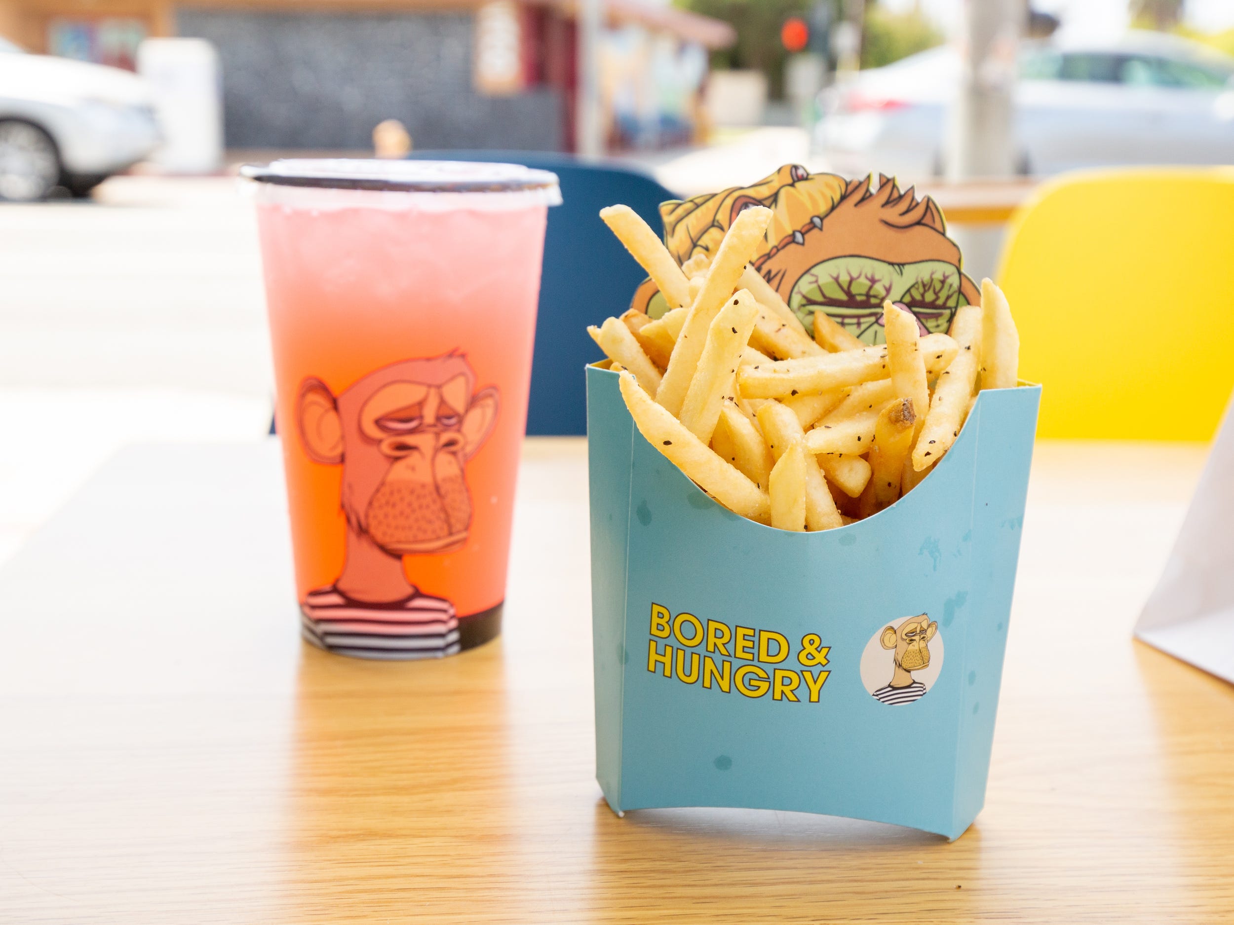 A pink beverage inside a plastic cup and fries in the foreground. The fries packaging has more ape branding.