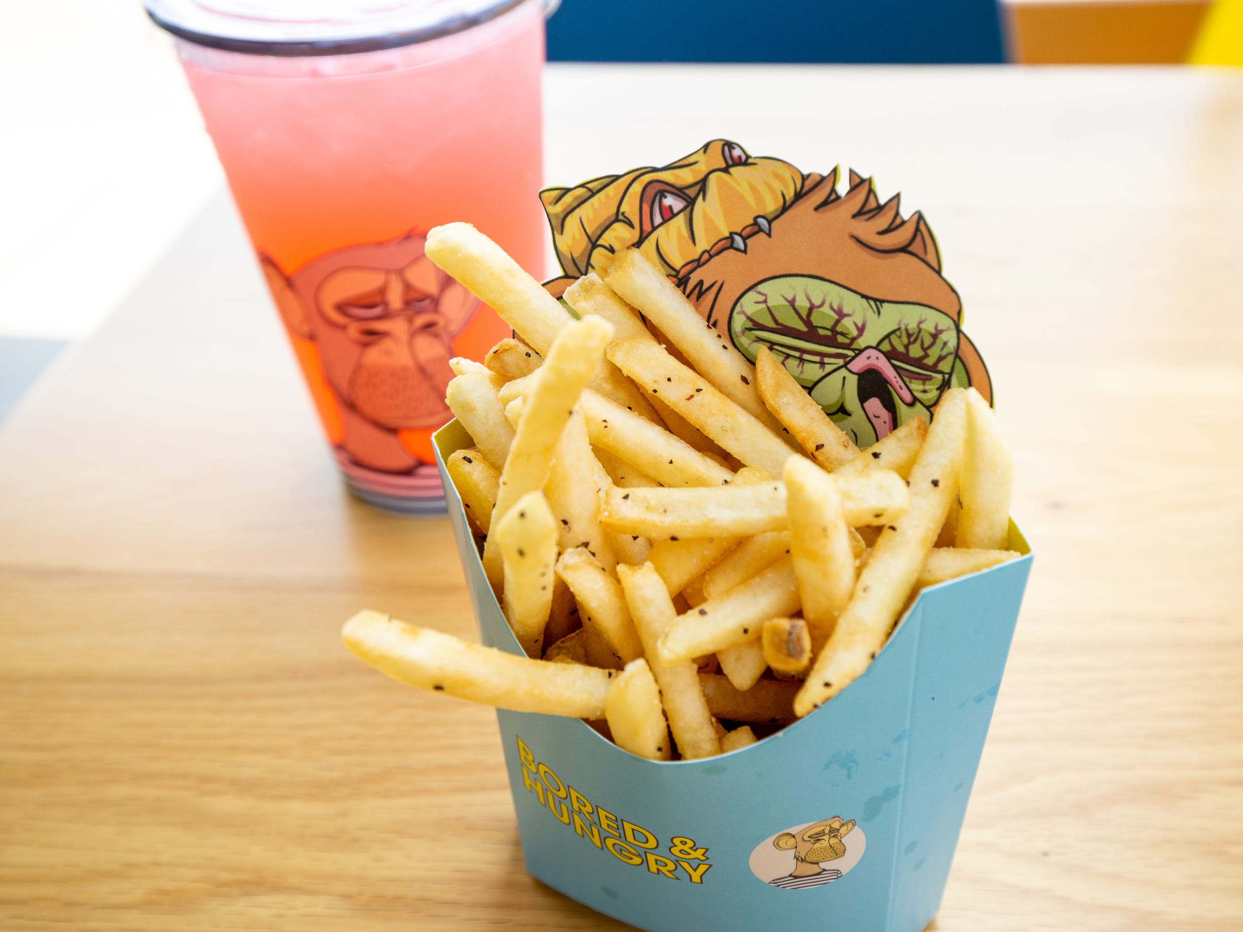 A pink beverage inside a plastic cup and fries in the foreground. The fries packaging has more ape branding.