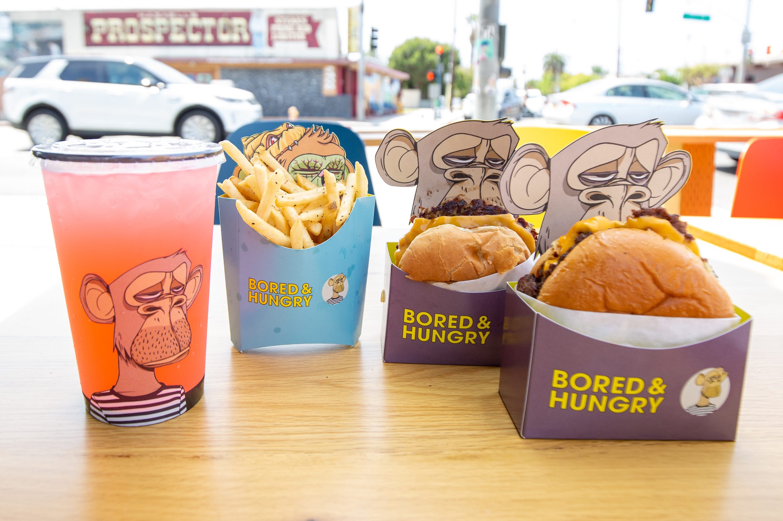 Two burgers, fries, a pink drink all in ape branded Bored and Hungry packaging.