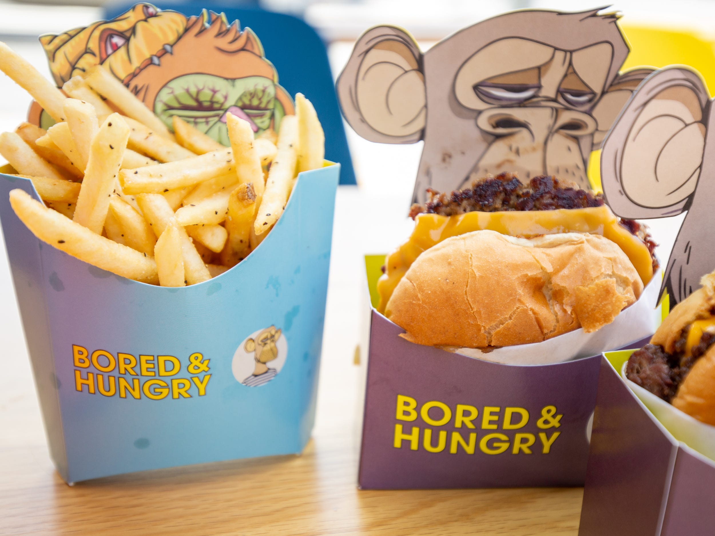 Fries inside of a Bored and Hungry container with an ape on the packaging. It's next to a cheeseburger with similar packaging.