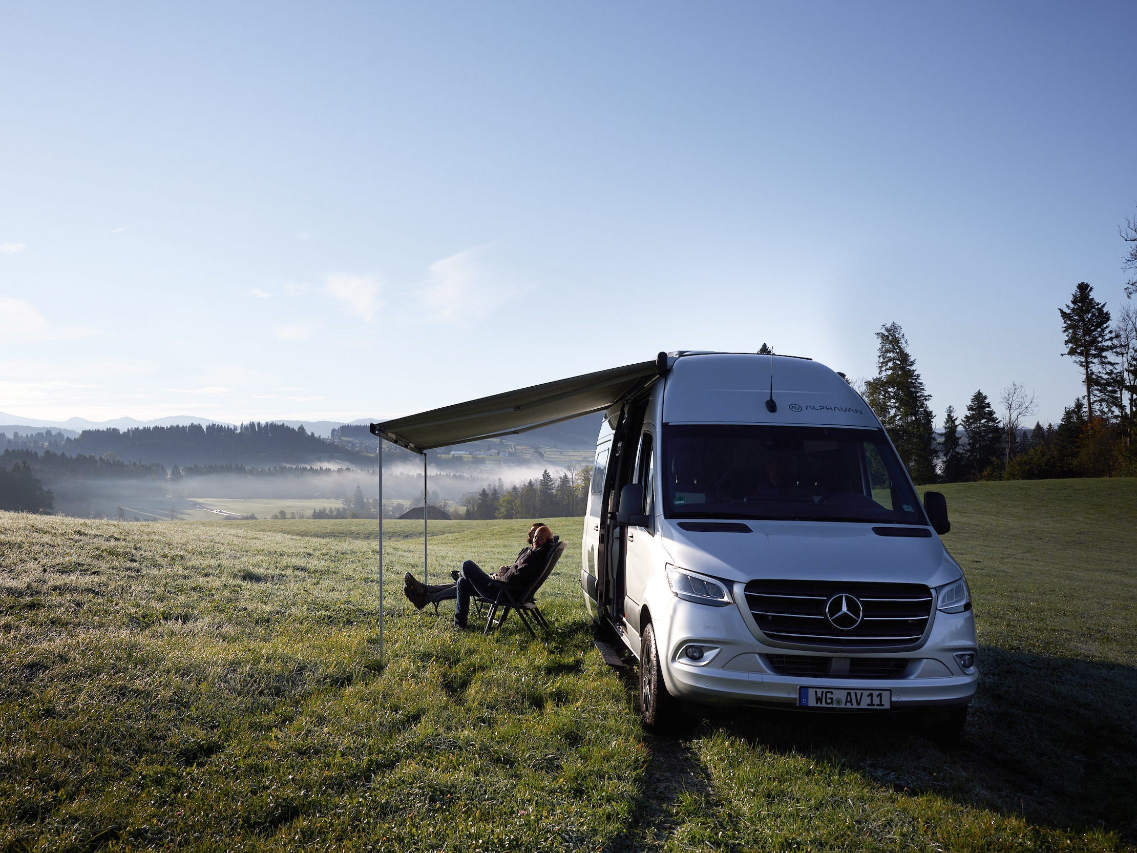 Alphavan's camper van in a grassy field with the awning out. Two people are sitting under the awning.