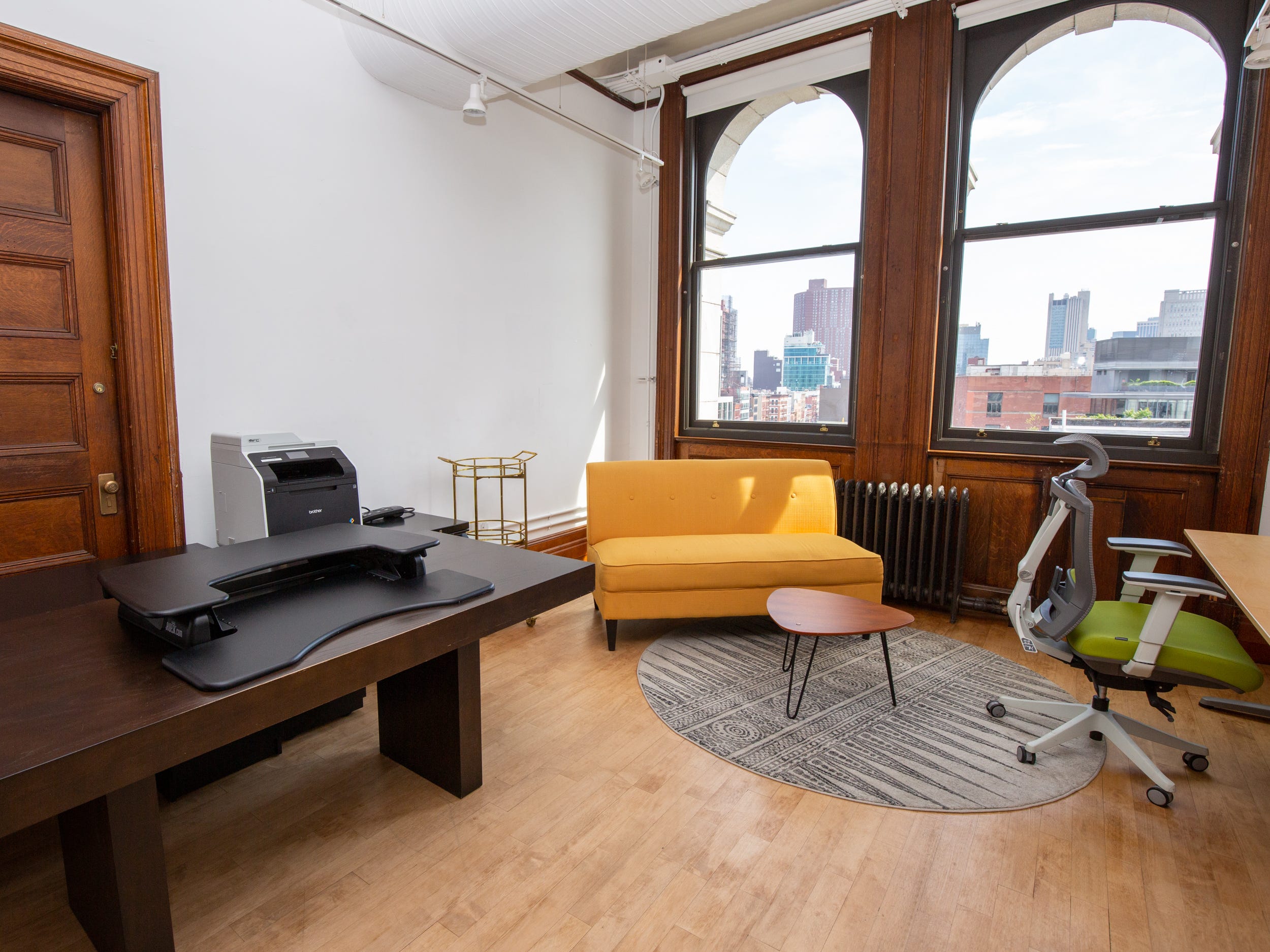 A room with a couch, long tables, office chair near windows.