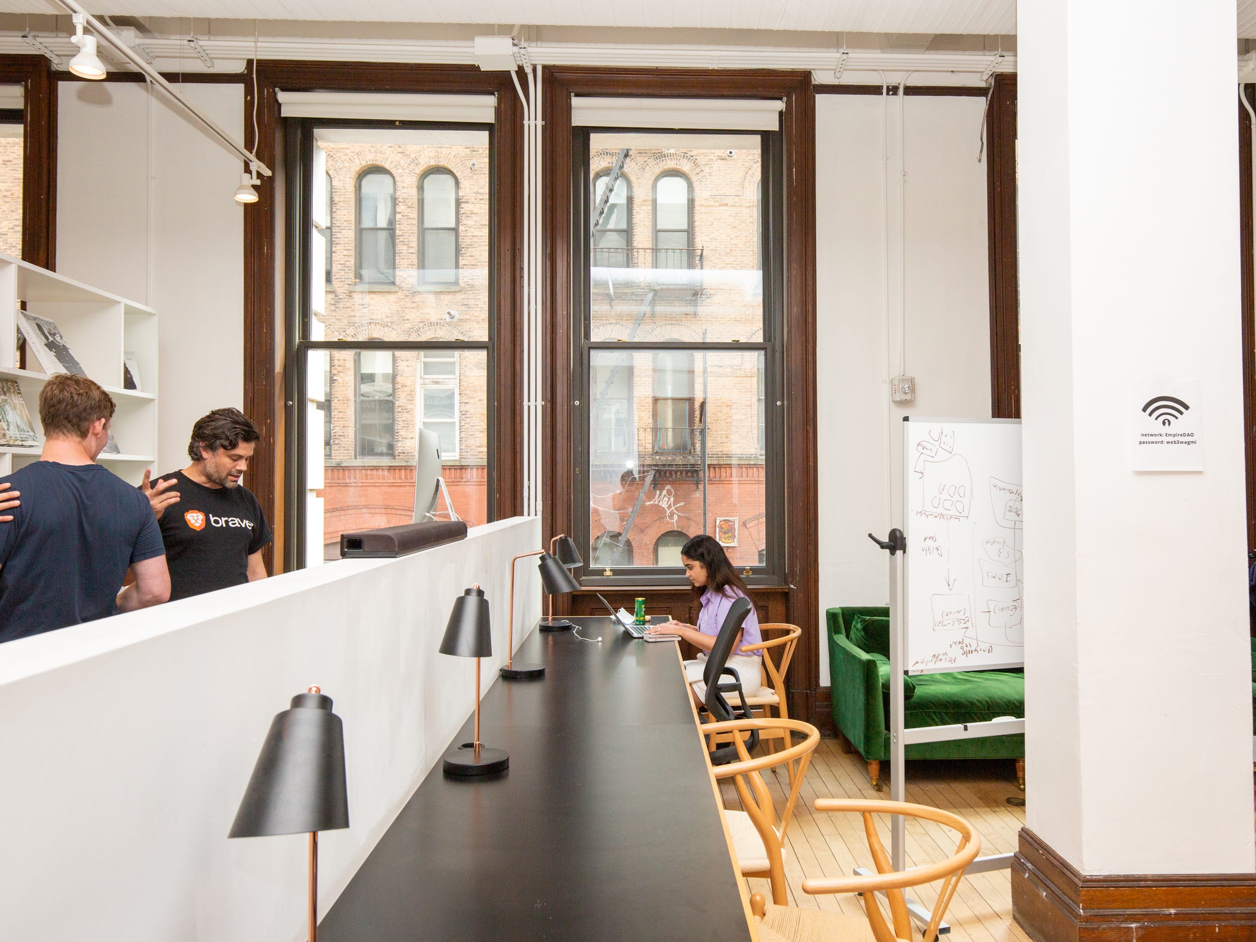 A long table with chairs, table lamps. Someone is working at the end of the table. People are standing nearby.