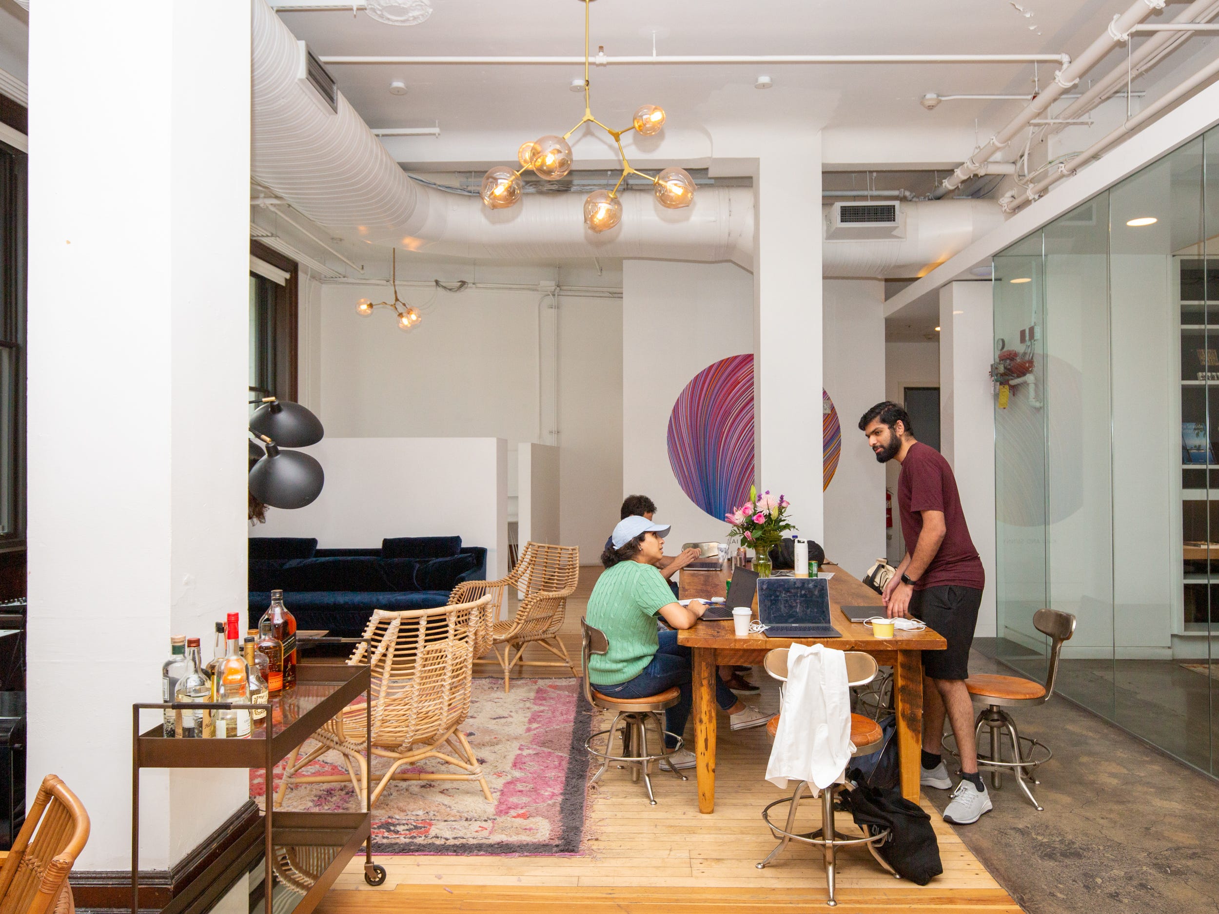 People are working at a long table nearby in a room with colorful rugs, chairs, bar cart, lights.