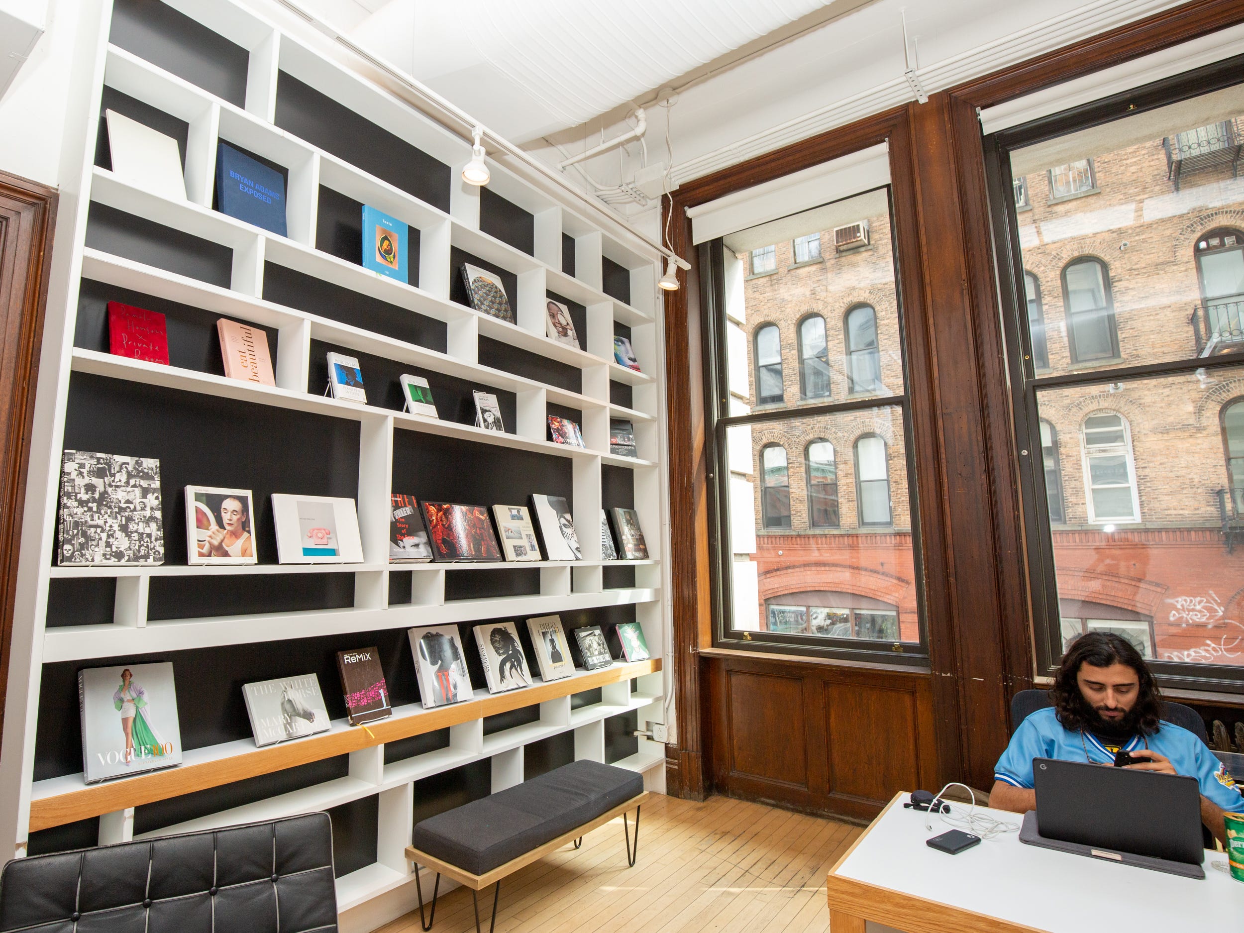 A room with someone working at a desk, large shelves filled with items, and large windows.