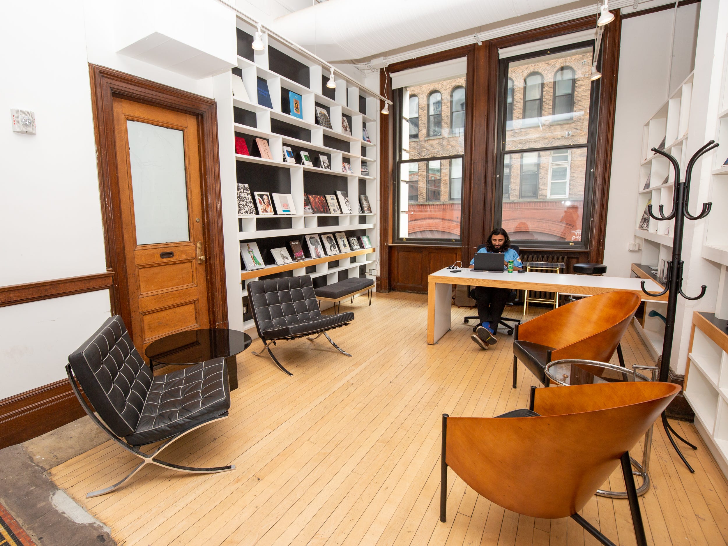 A room with lounge chairs, someone working at a desk, large shelves filled with items, and large windows.