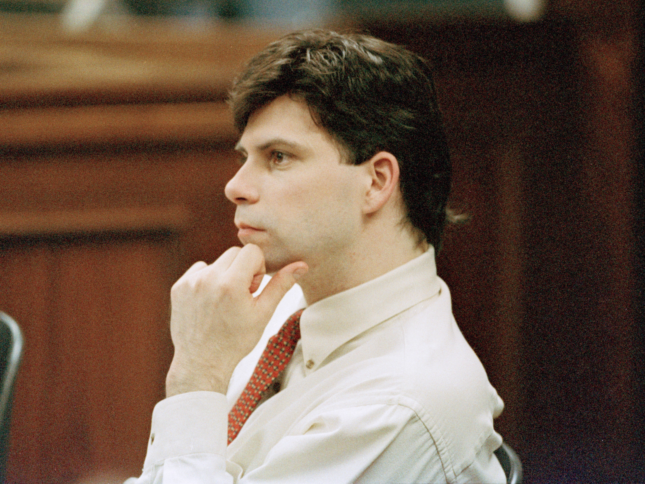 A man with dark hair in a white shirt and a red tie sitting in a court room.